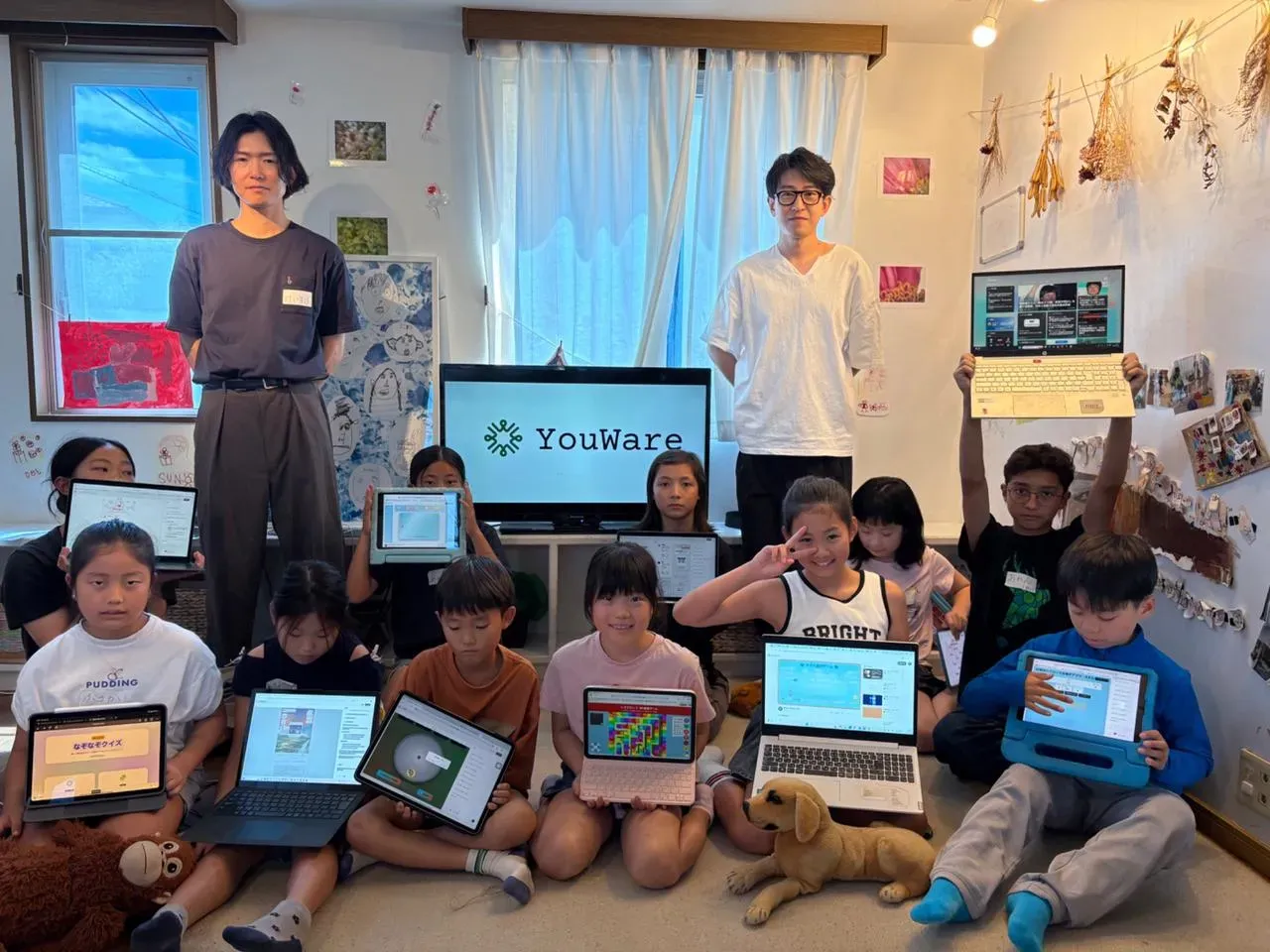 A group of children in a Tokyo classroom holding tablets and laptops during a creative coding workshop